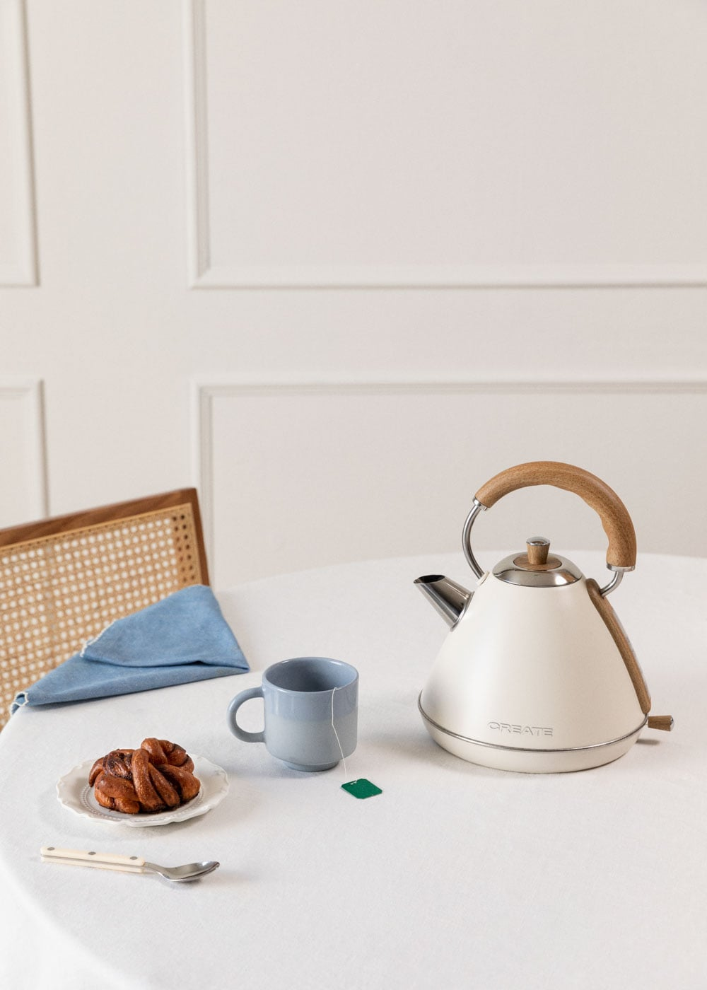 Un hervidor blanco con asa de madera, una taza azul y un bollo sobre una mesa.