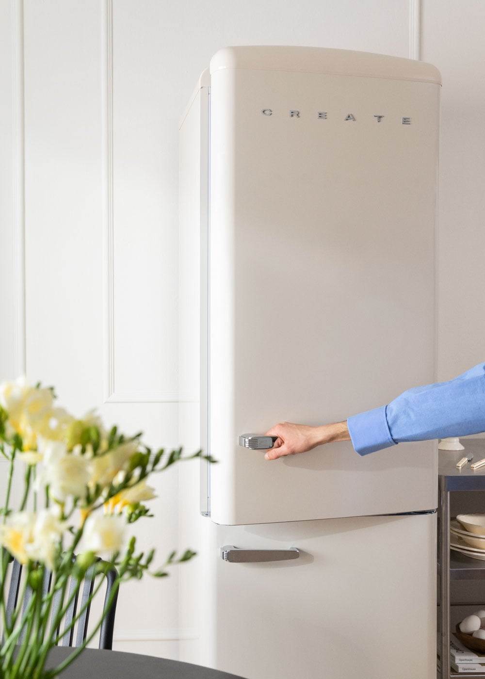 Una mano abre un refrigerador color crema con tiradores plateados, junto a flores.