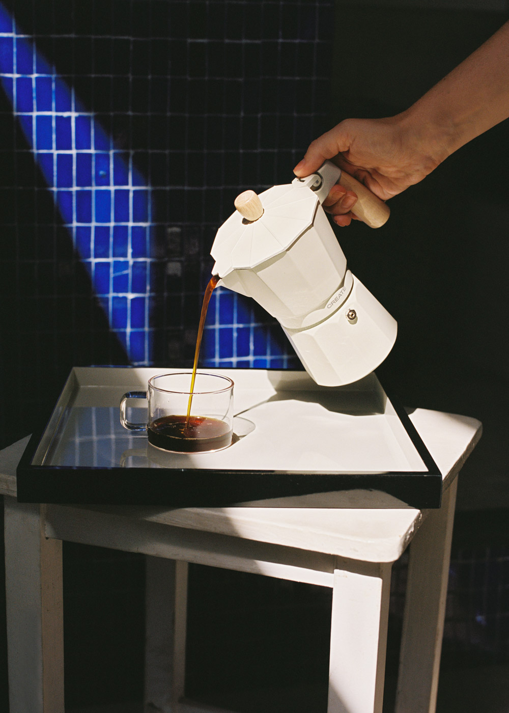 Una cafetera blanca vierte café en una taza de cristal sobre una bandeja.