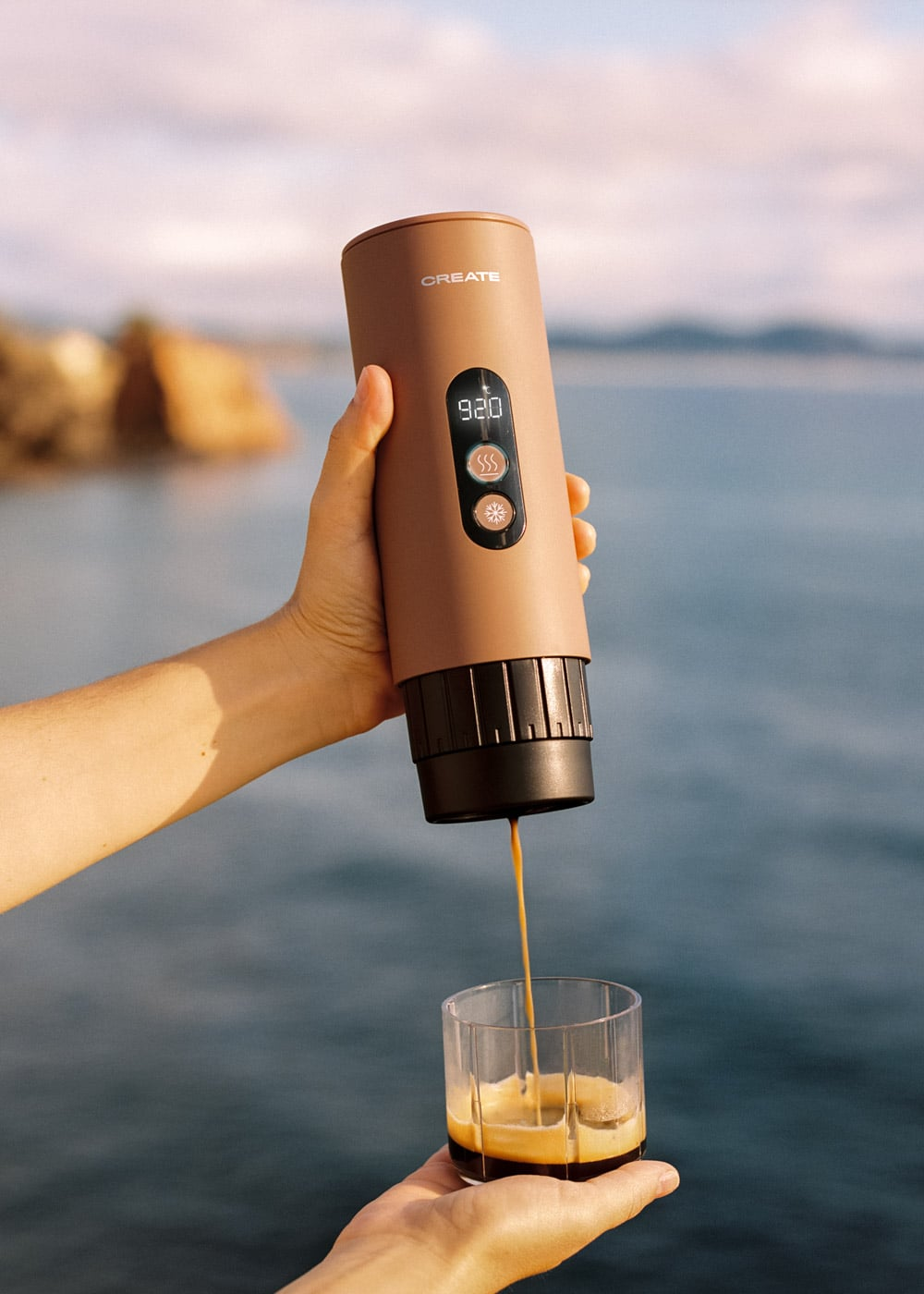 Una cafetera marrón vierte espresso en un vaso junto al mar.