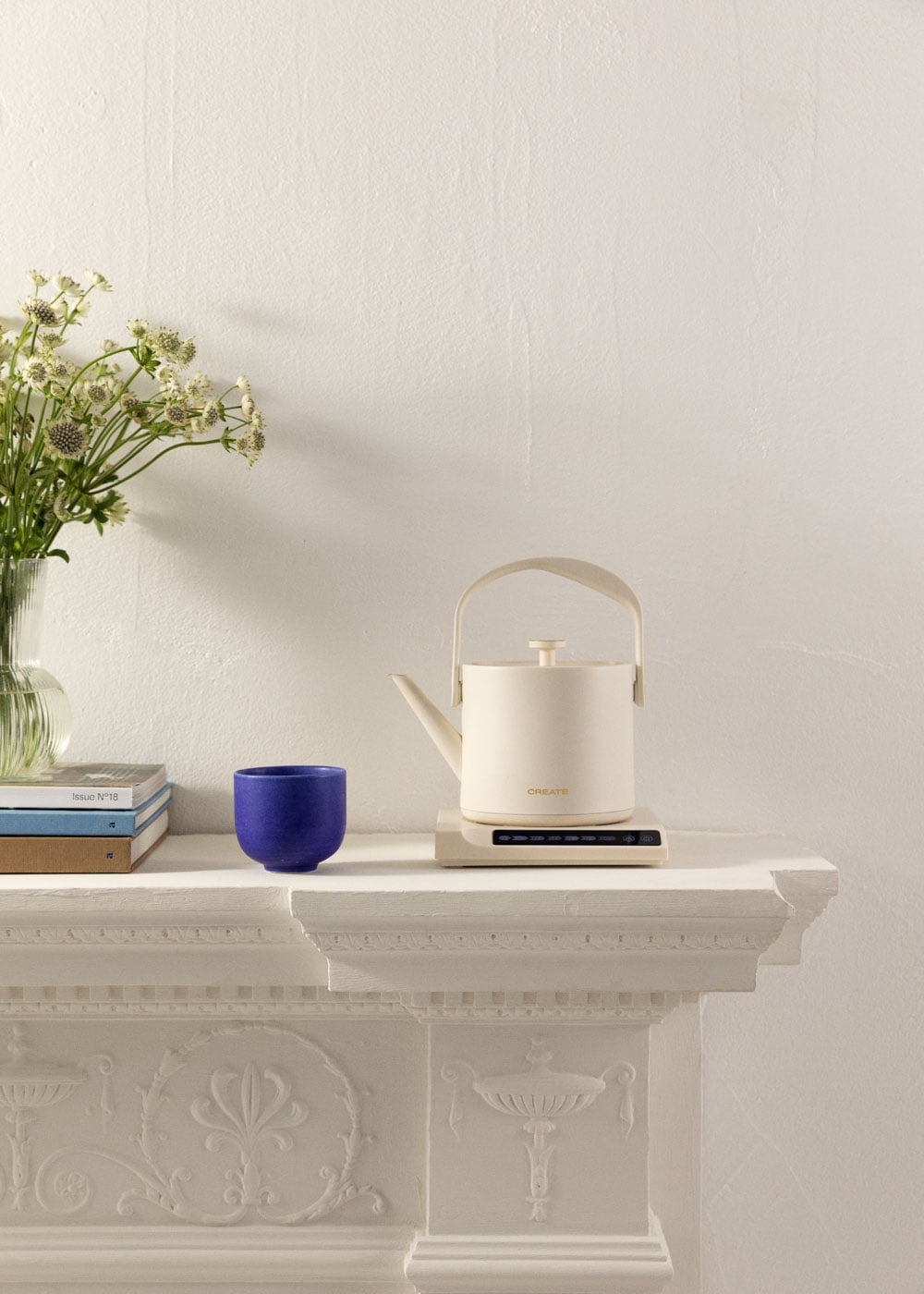 Una tetera color crema se asienta sobre una base junto a una taza azul y flores.