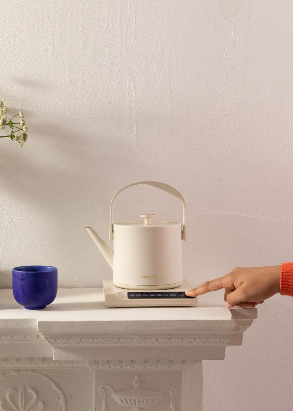 Una mano presiona un botón en la base de un hervidor beige con una taza azul cerca.