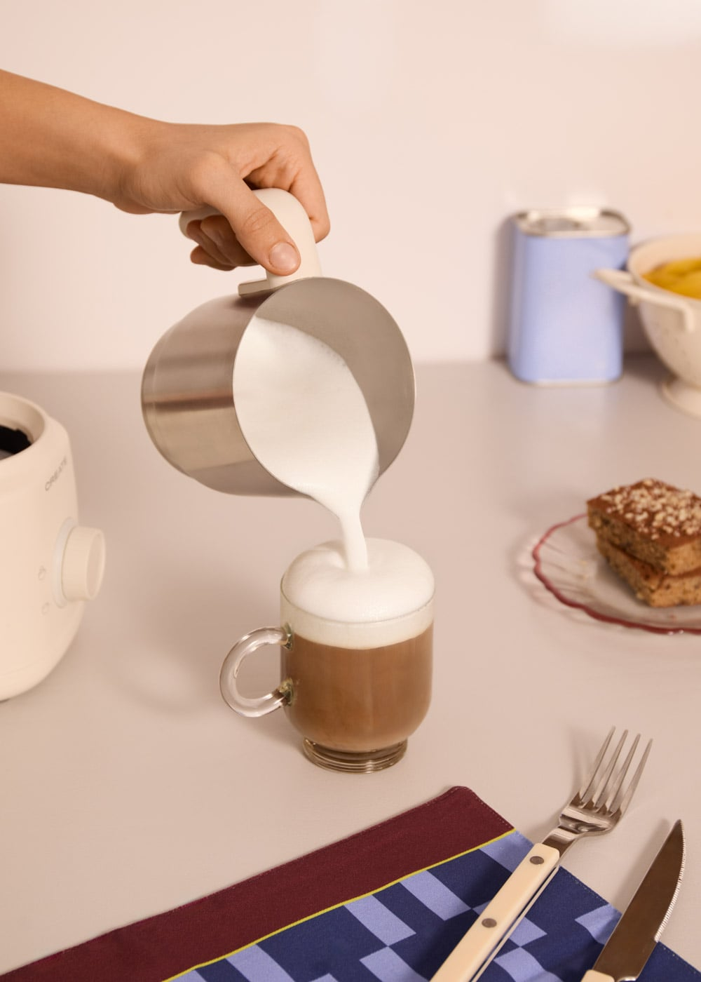 Una mano vierte leche espumosa de una jarra de metal en una taza de café de cristal.