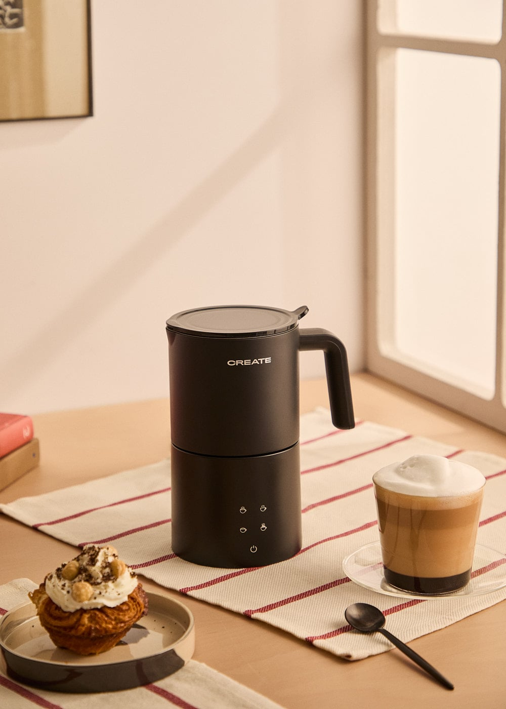 Un espumador de leche negro junto a un pastel y un vaso de café espumoso.