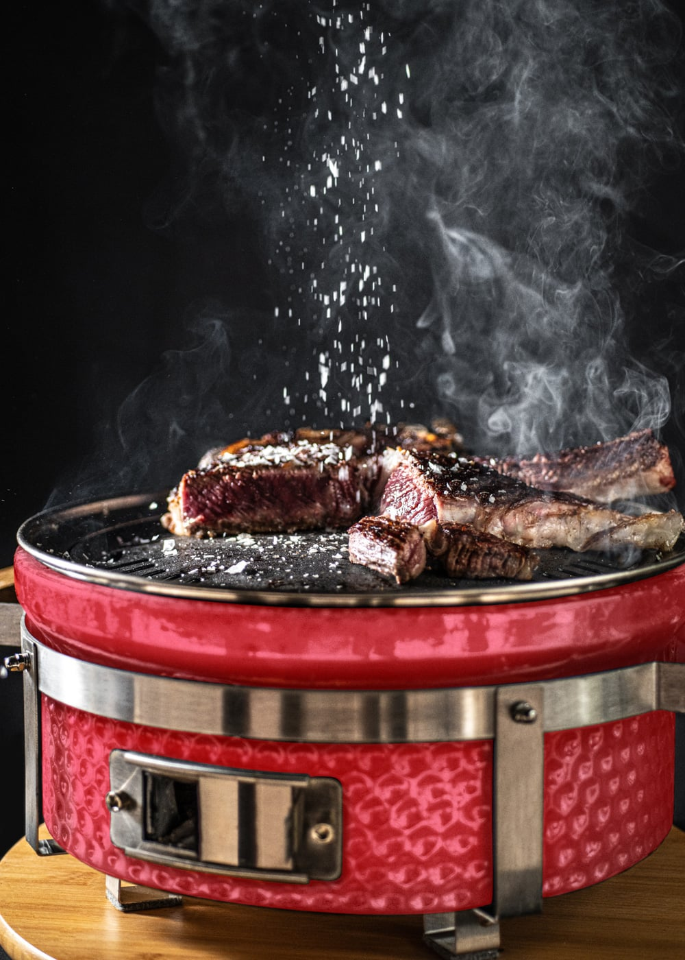 Un jugoso filete se sazona con sal en una parrilla roja, con humo ascendiendo.