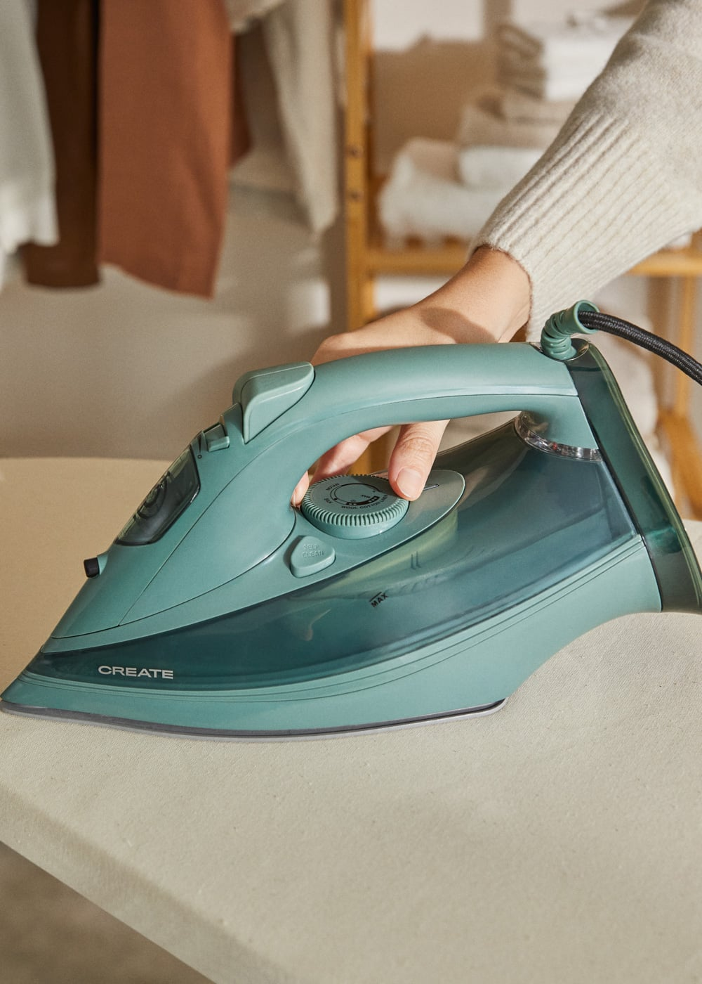 Una persona maneja una plancha de vapor turquesa sobre una tabla de planchar. Ropa colgada al fondo.