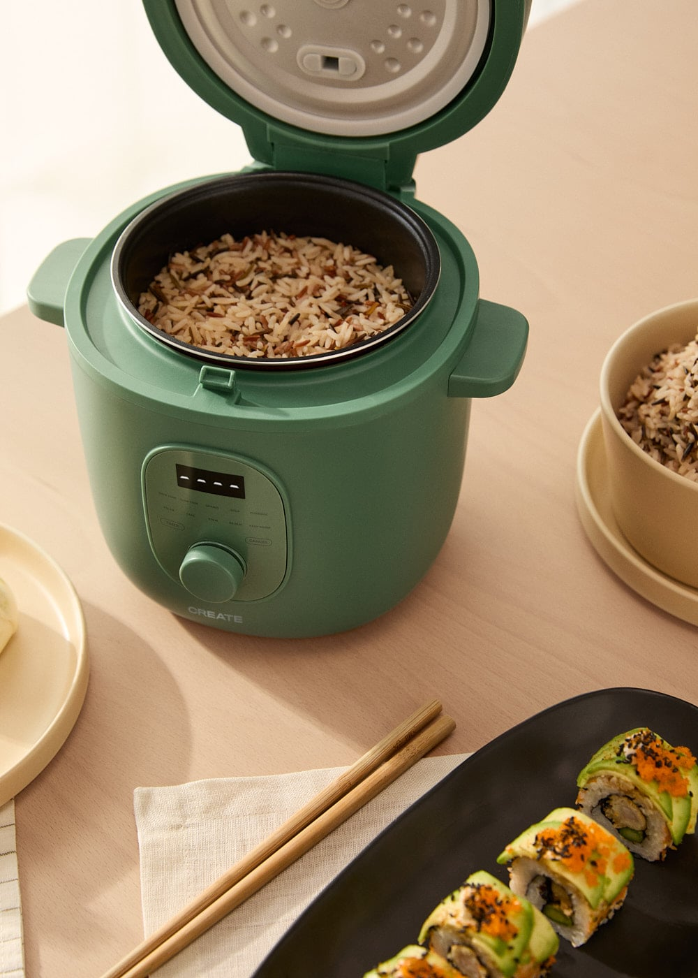 Una arrocera verde con tapa abierta, llena de arroz. Sushi y palillos cerca.