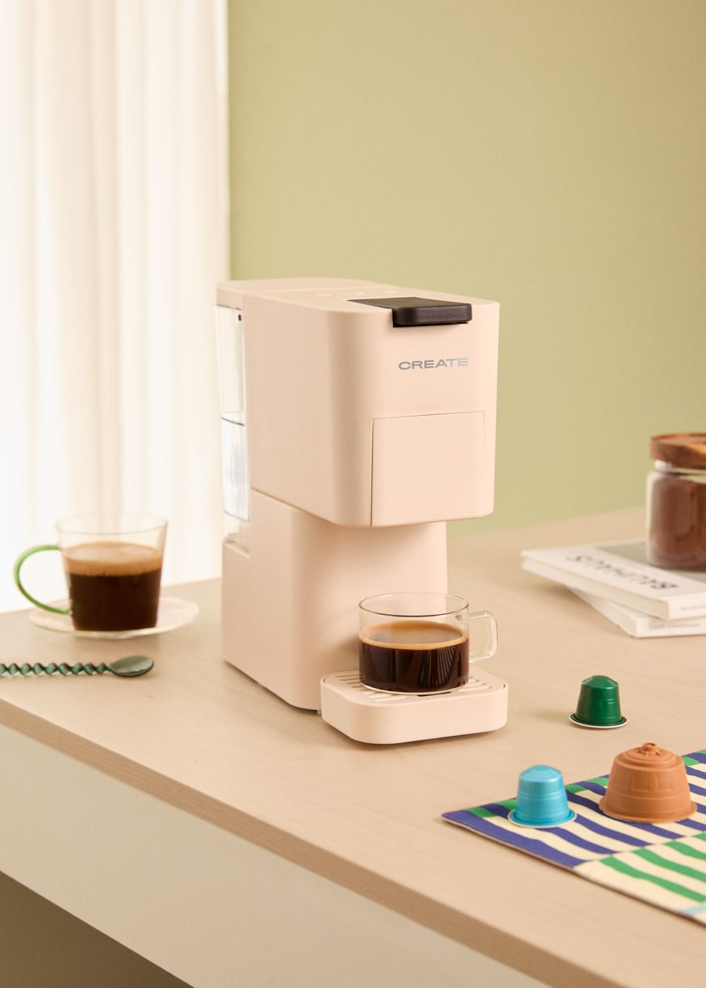 Una cafetera color crema prepara café en una taza pequeña. Hay varias cápsulas de café cerca.