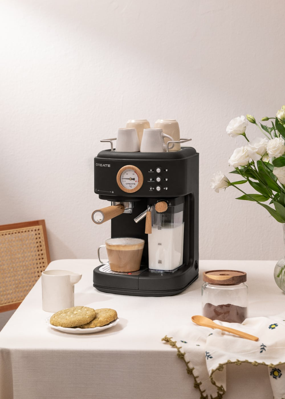 Una cafetera negra con tazas, jarra de leche, galletas y cacao en polvo.