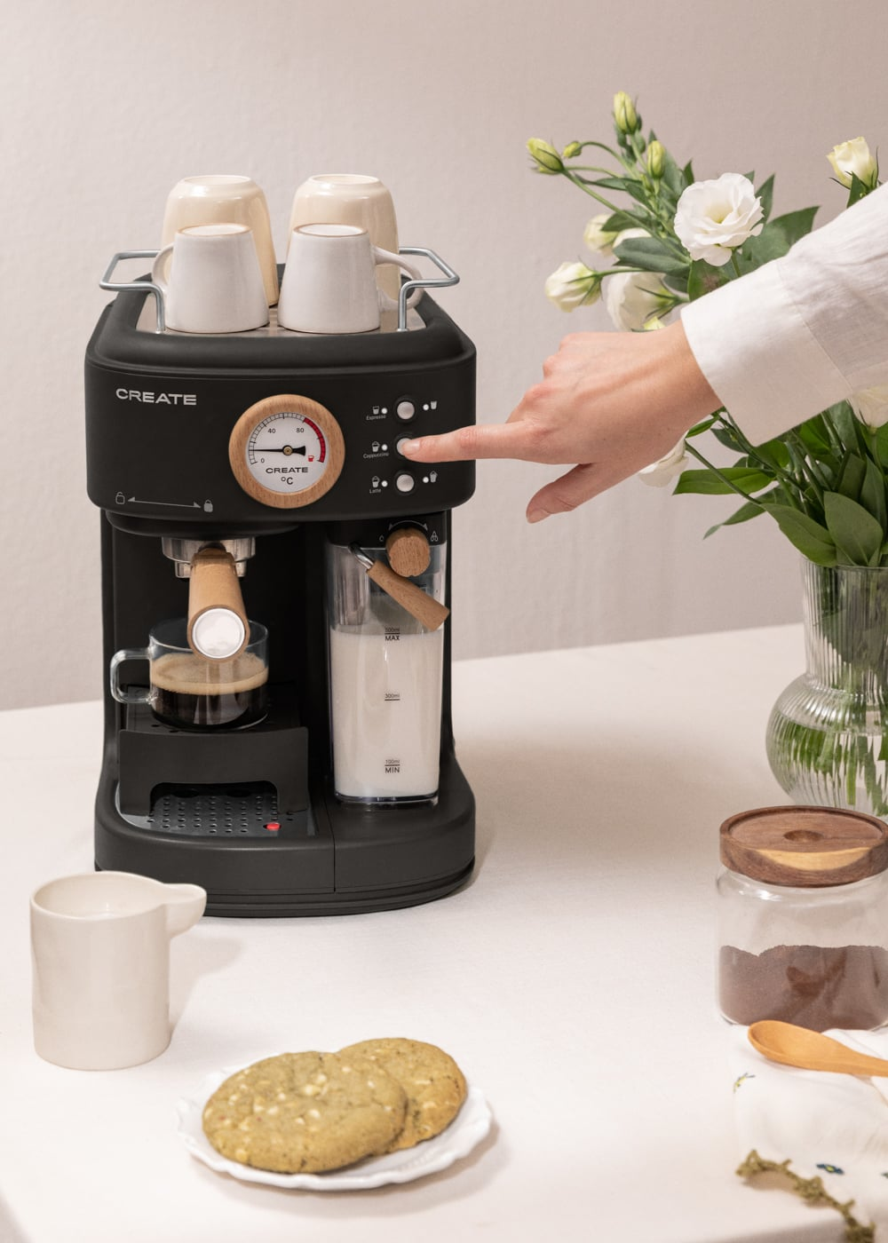 Una cafetera espresso negra con una mano presionando un botón, tazas encima y café preparándose.