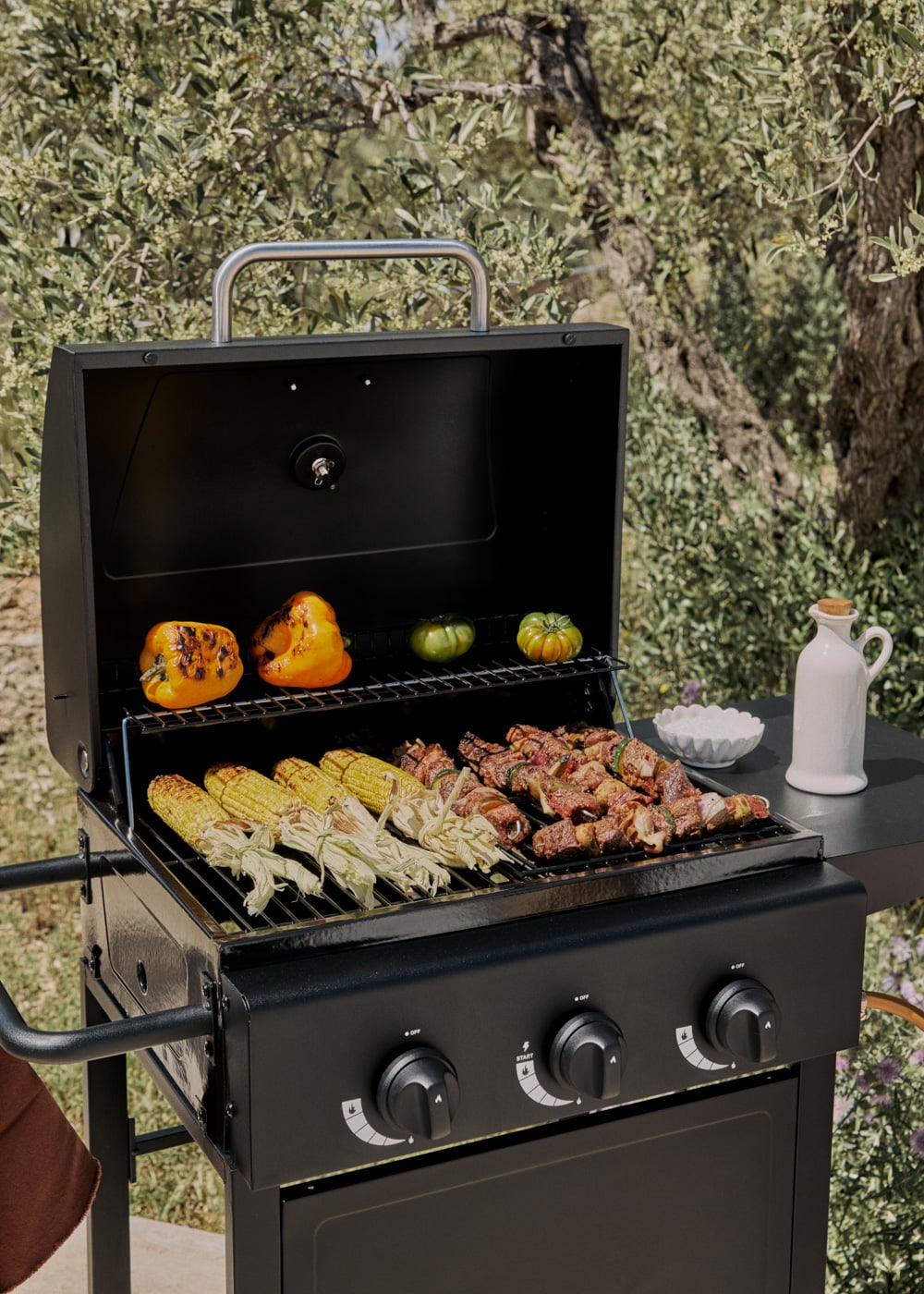 Una parrilla negra con maíz, brochetas de carne y pimientos asándose. Árboles al fondo.