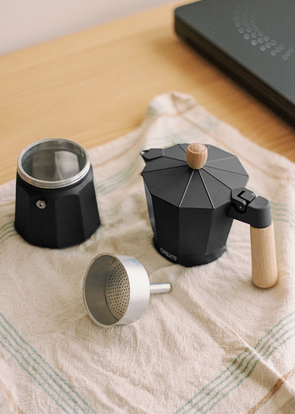 Cafetera moka negra con mango de madera y filtro sobre tela a cuadros.