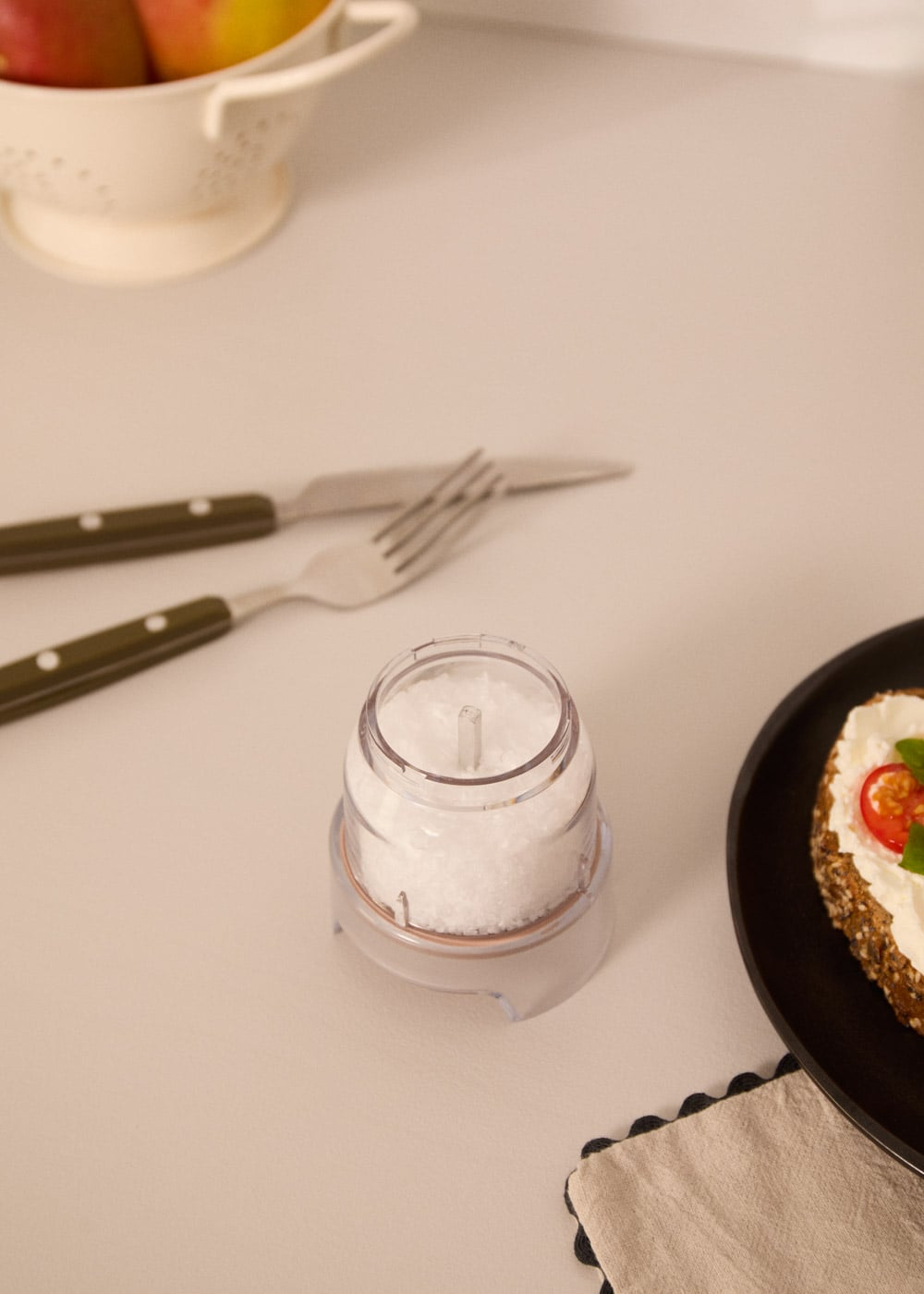Un molinillo de sal transparente está sobre una mesa con un tenedor, cuchillo y una rebanada de tostada.
