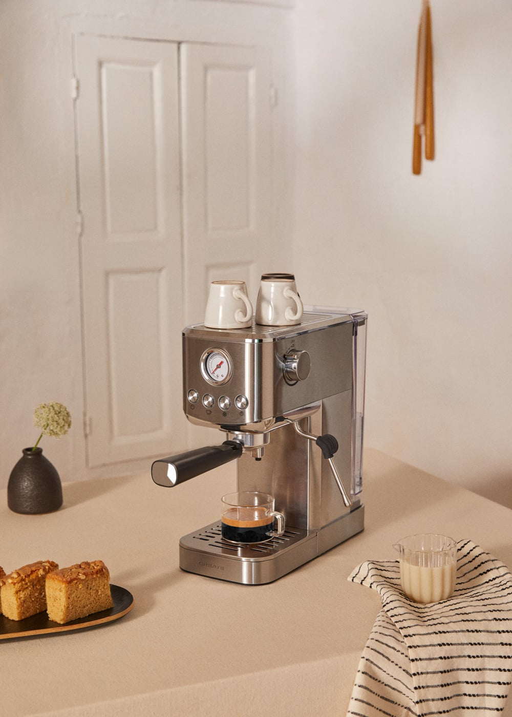 Máquina de espresso con dos tazas encima. Al lado, un vaso de espresso y pastel.