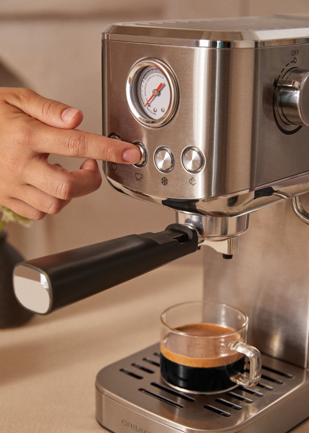 Una mano presiona un botón en una máquina de espresso plateada que prepara café en una taza de cristal.