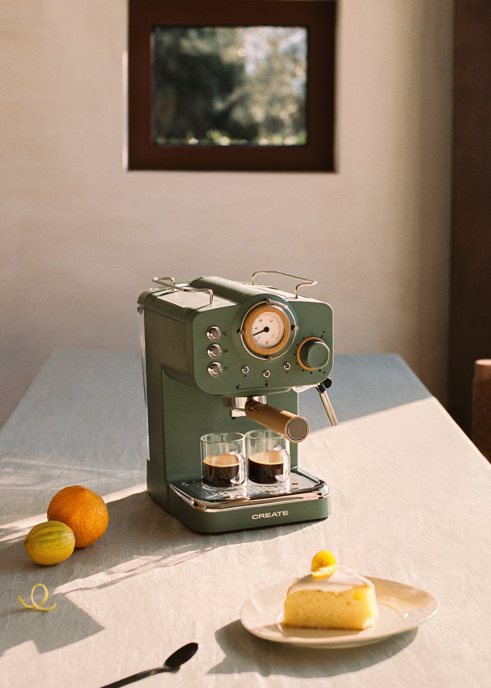 Una máquina de espresso verde con dos vasos de café, naranjas y un trozo de pastel.