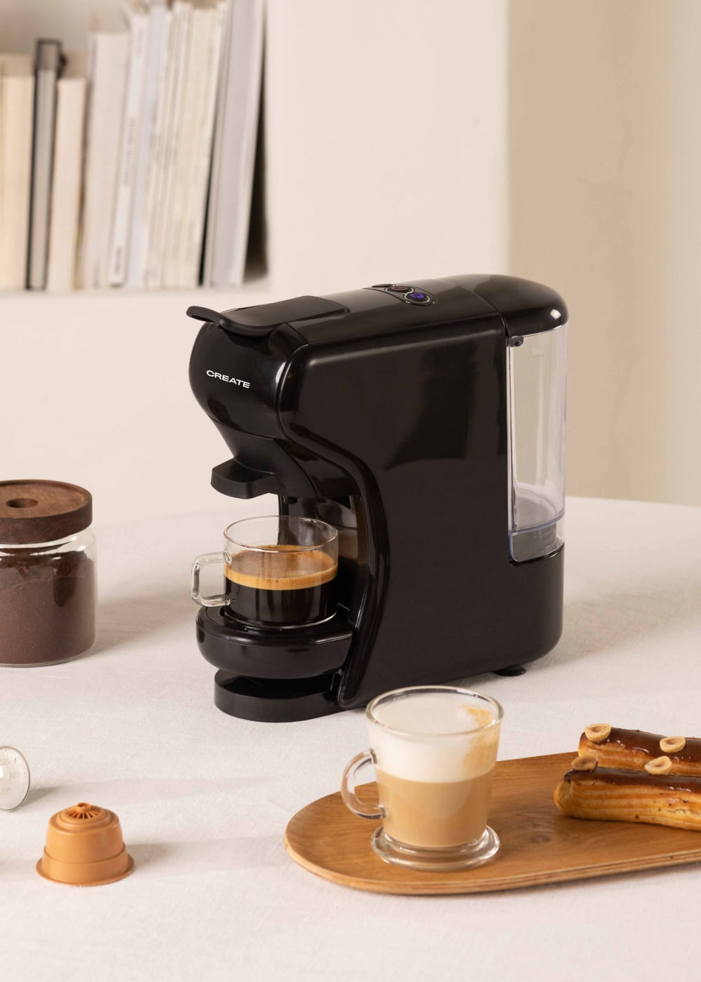 Cafetera negra con espresso, café con leche y éclairs sobre mesa.