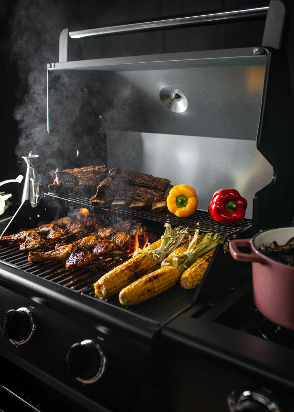 Costillas y maíz asándose en una barbacoa con pimientos y una olla rosa.