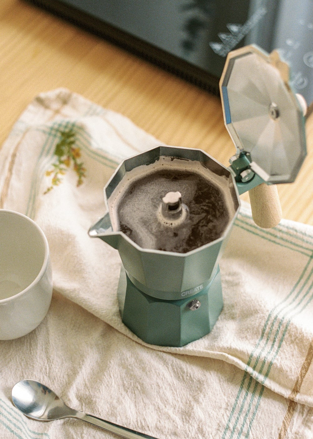 Una cafetera moka turquesa prepara café sobre un paño a rayas, con cuchara y taza cerca.