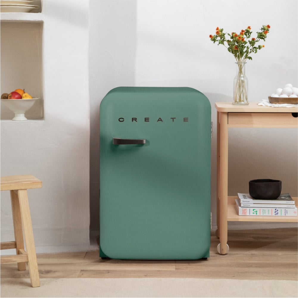 Un refrigerador verde con la palabra CREATE junto a una mesa de madera con huevos y flores.
