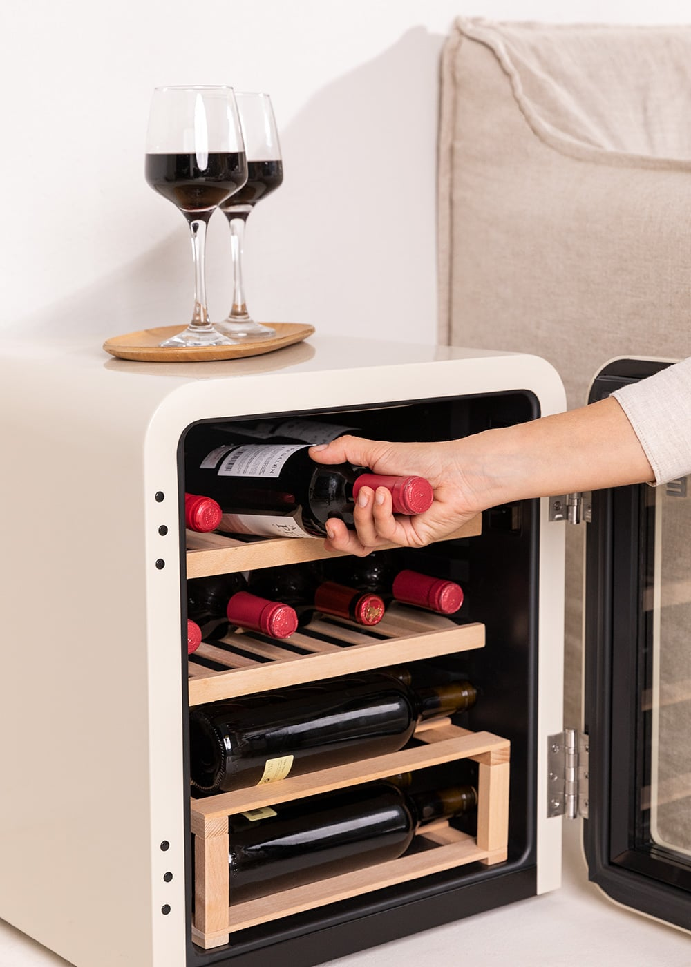 Una mano saca una botella de vino de una nevera color crema. Dos copas de vino tinto están al lado.