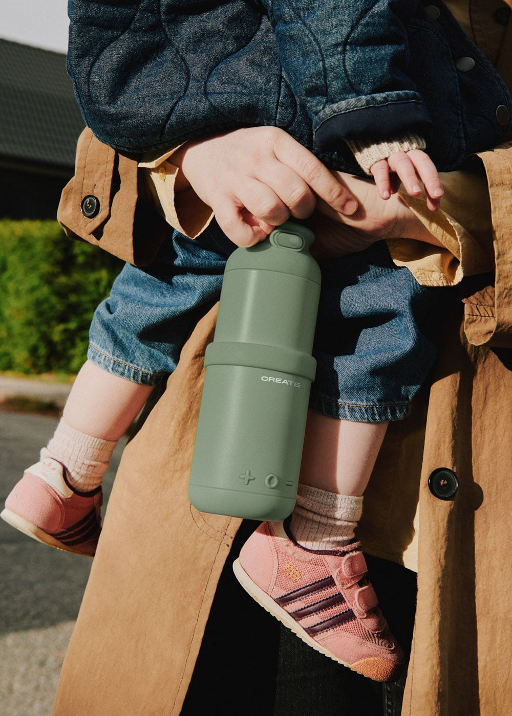 Un niño con zapatos rosas sostiene una botella verde. Una persona lleva vaqueros.