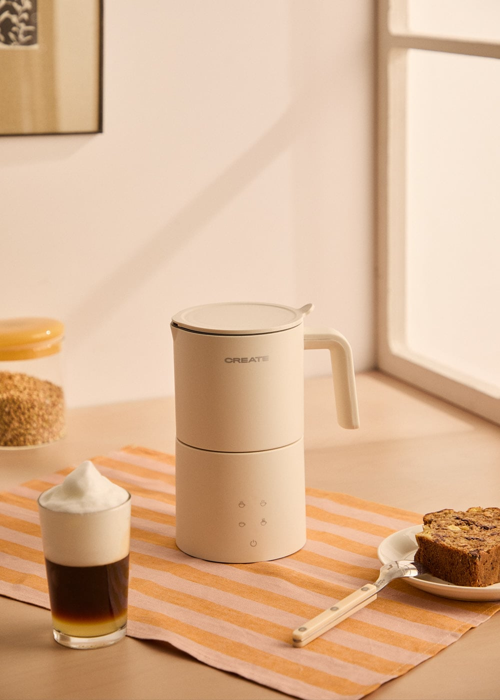 Una cafetera beige sobre un paño a rayas con un vaso de café espumoso y pastel.