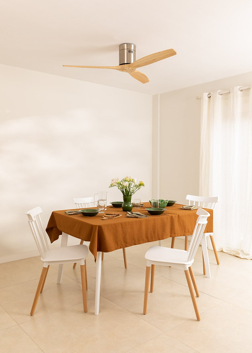 Mesa de comedor con mantel marrón, cuatro sillas blancas, vajilla y flores.