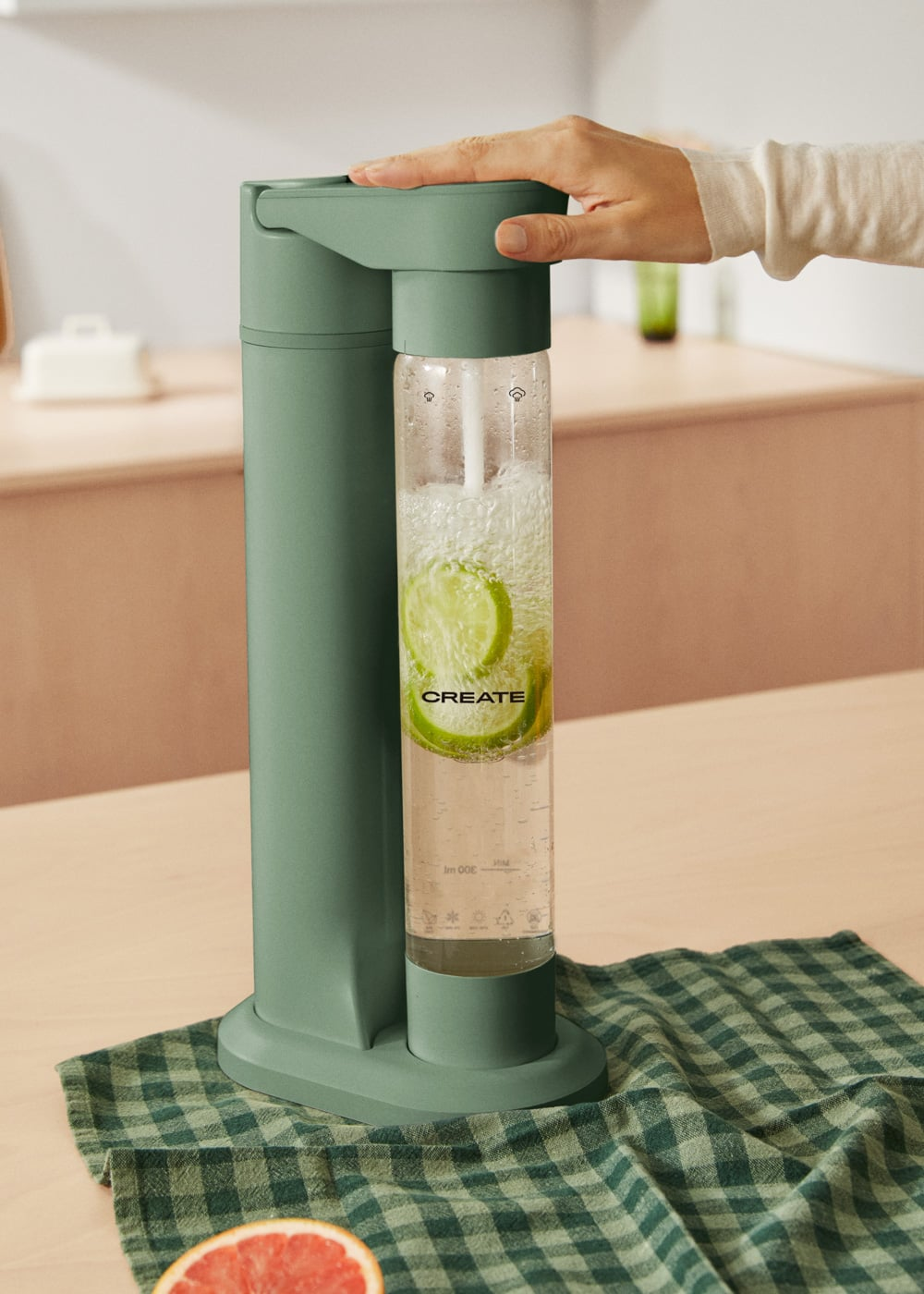 Una mano presiona una máquina de soda verde con rodajas de lima en la botella. Un paño a cuadros debajo.