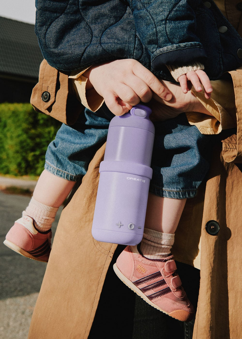 Un niño sostiene una botella de agua morada. Viste jeans y zapatos rosas.