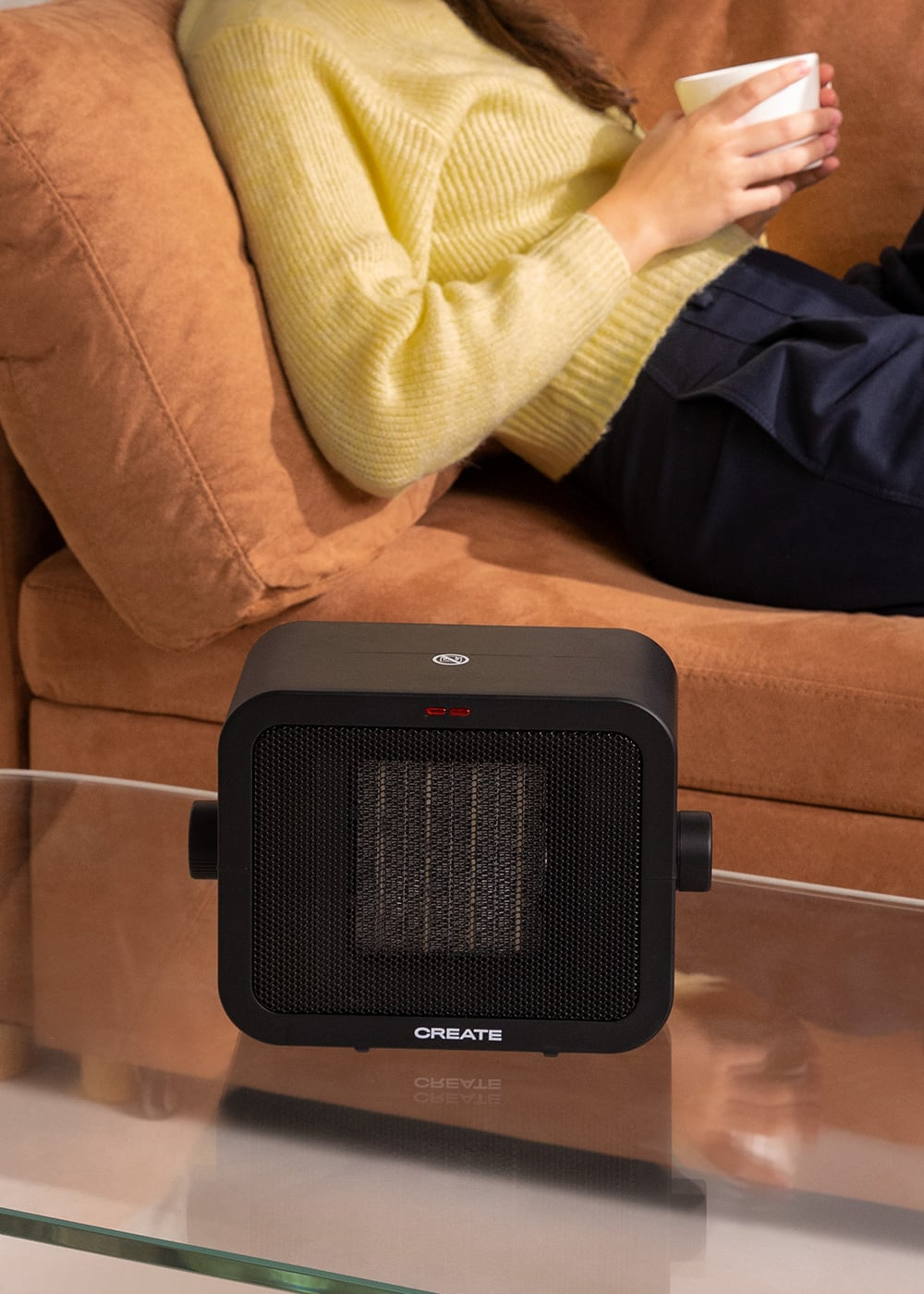 Un calefactor negro está sobre una mesa de cristal frente a un sofá marrón donde está sentada una persona.