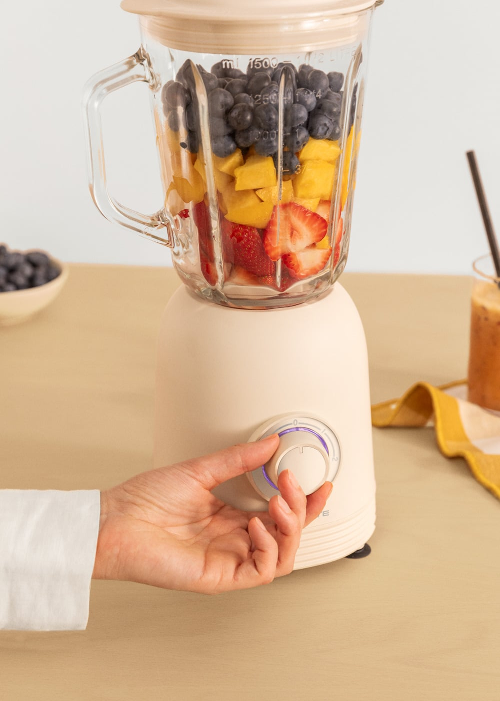 Una mano gira el dial de una licuadora color crema con bayas, mango y fresas.