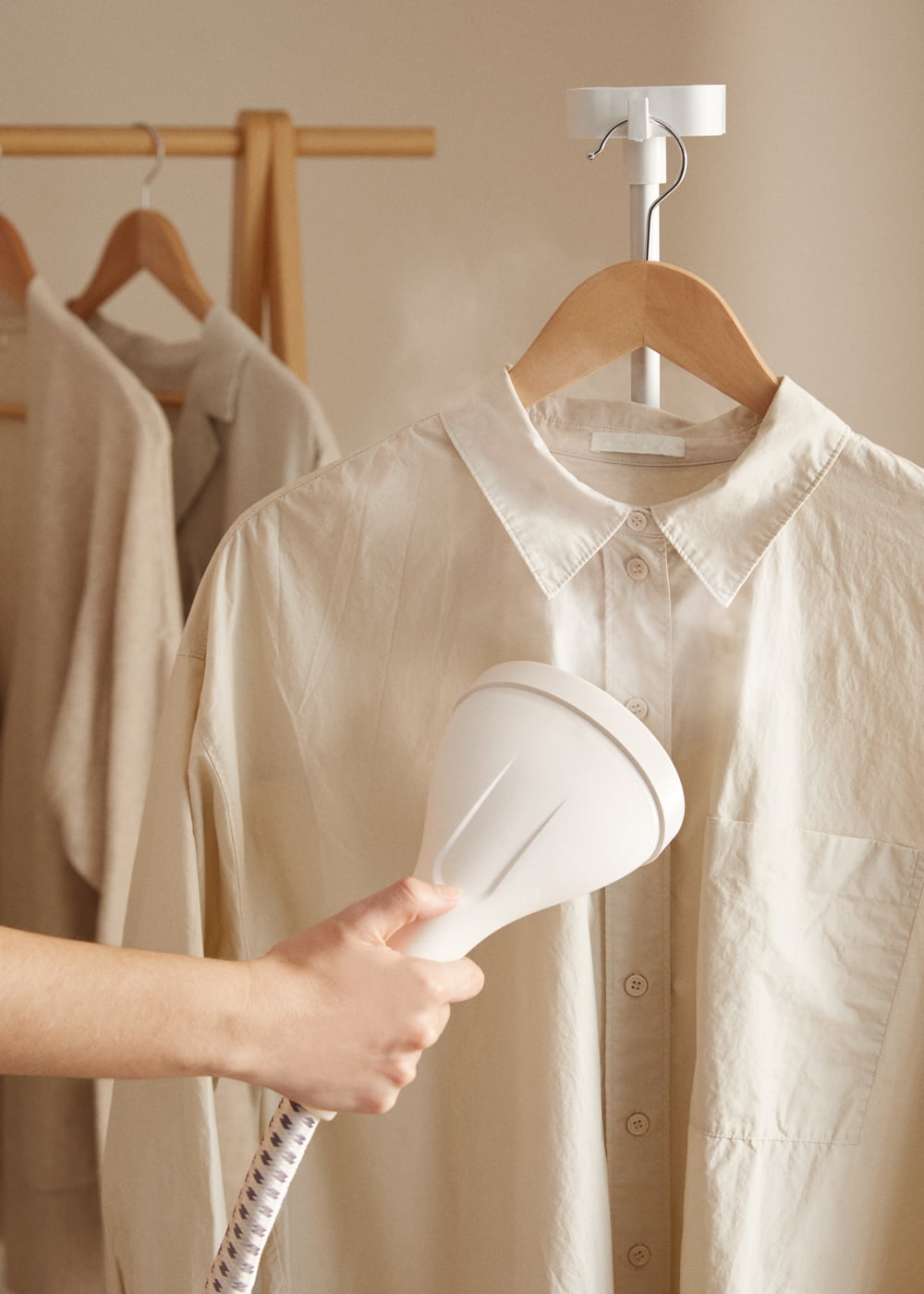 Una mano usa un vaporizador de ropa para alisar una camisa beige claro en una percha.