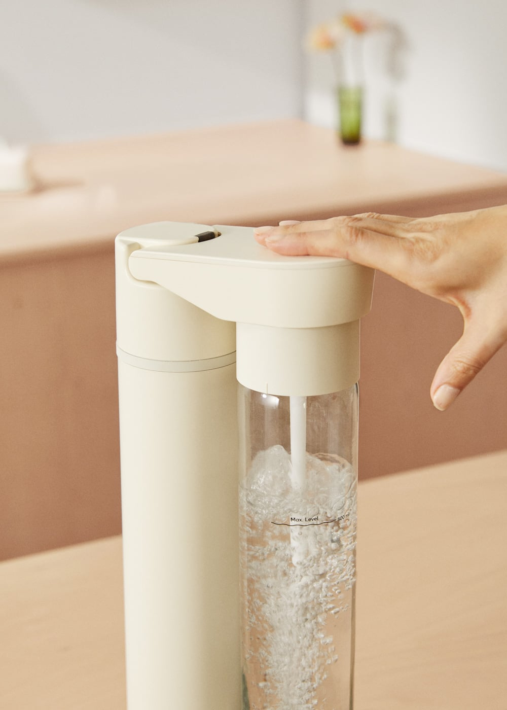 Una mano presiona un carbonatador de agua color crema con una botella llena.