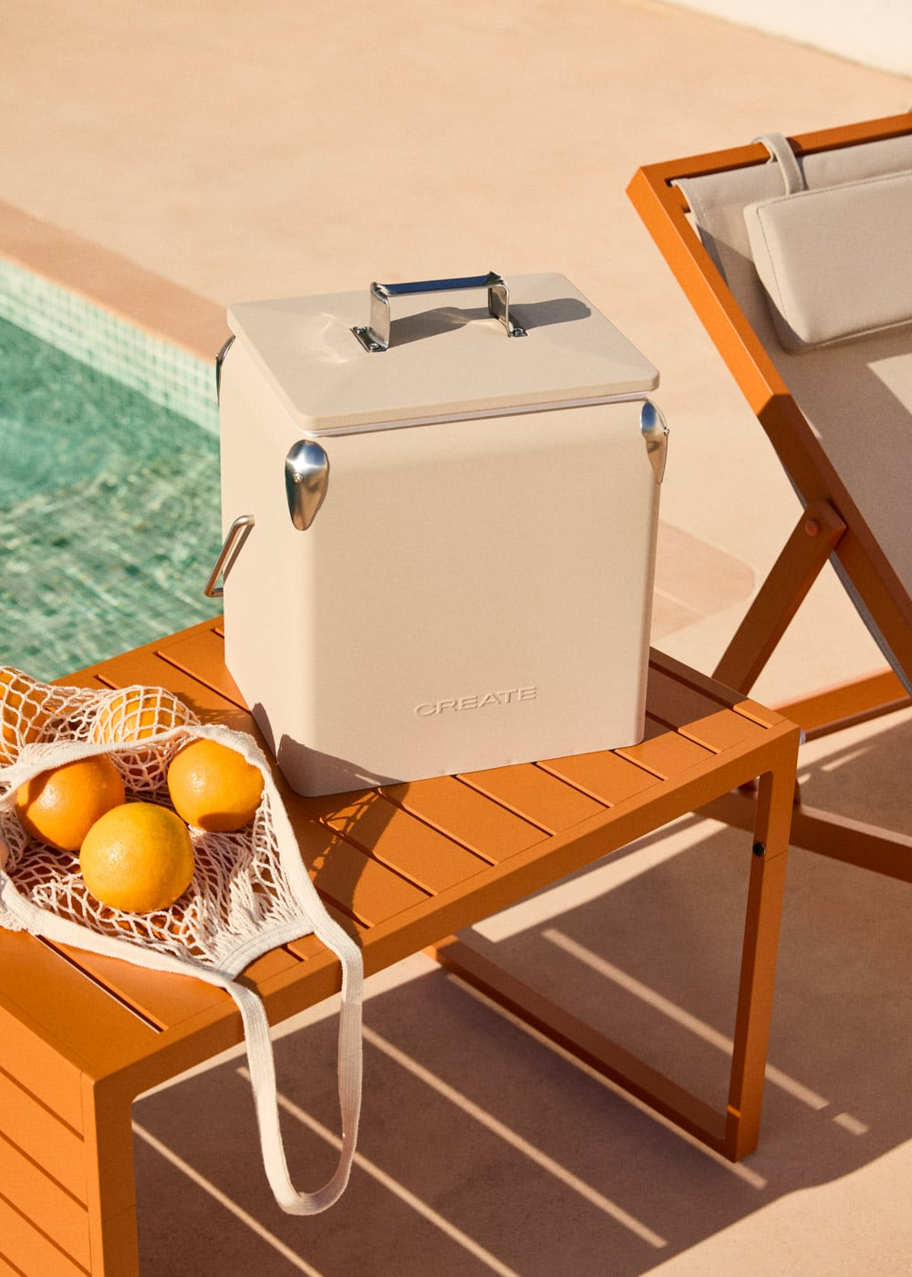 Una hielera beige sobre una mesa naranja junto a una piscina. Naranjas en una red.