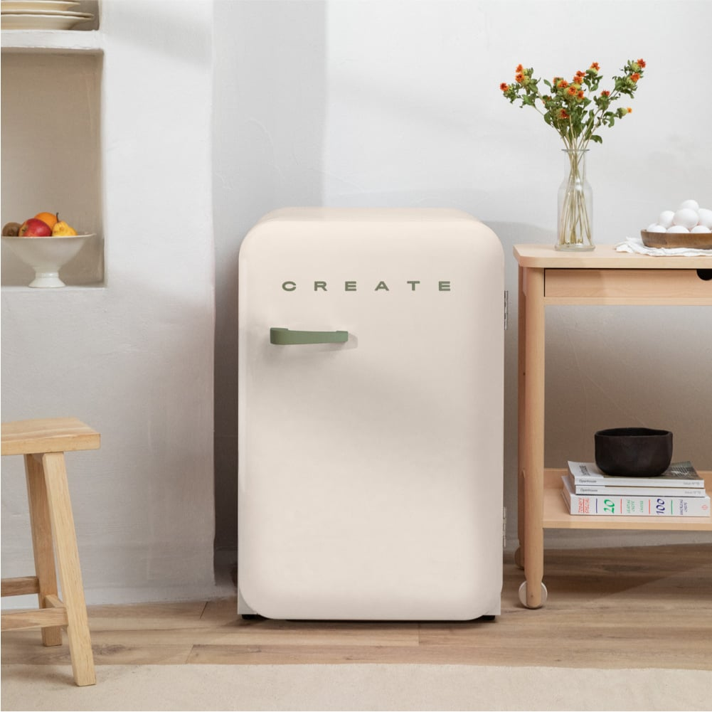 Un refrigerador color crema con asa verde junto a una mesa de madera con huevos y flores.