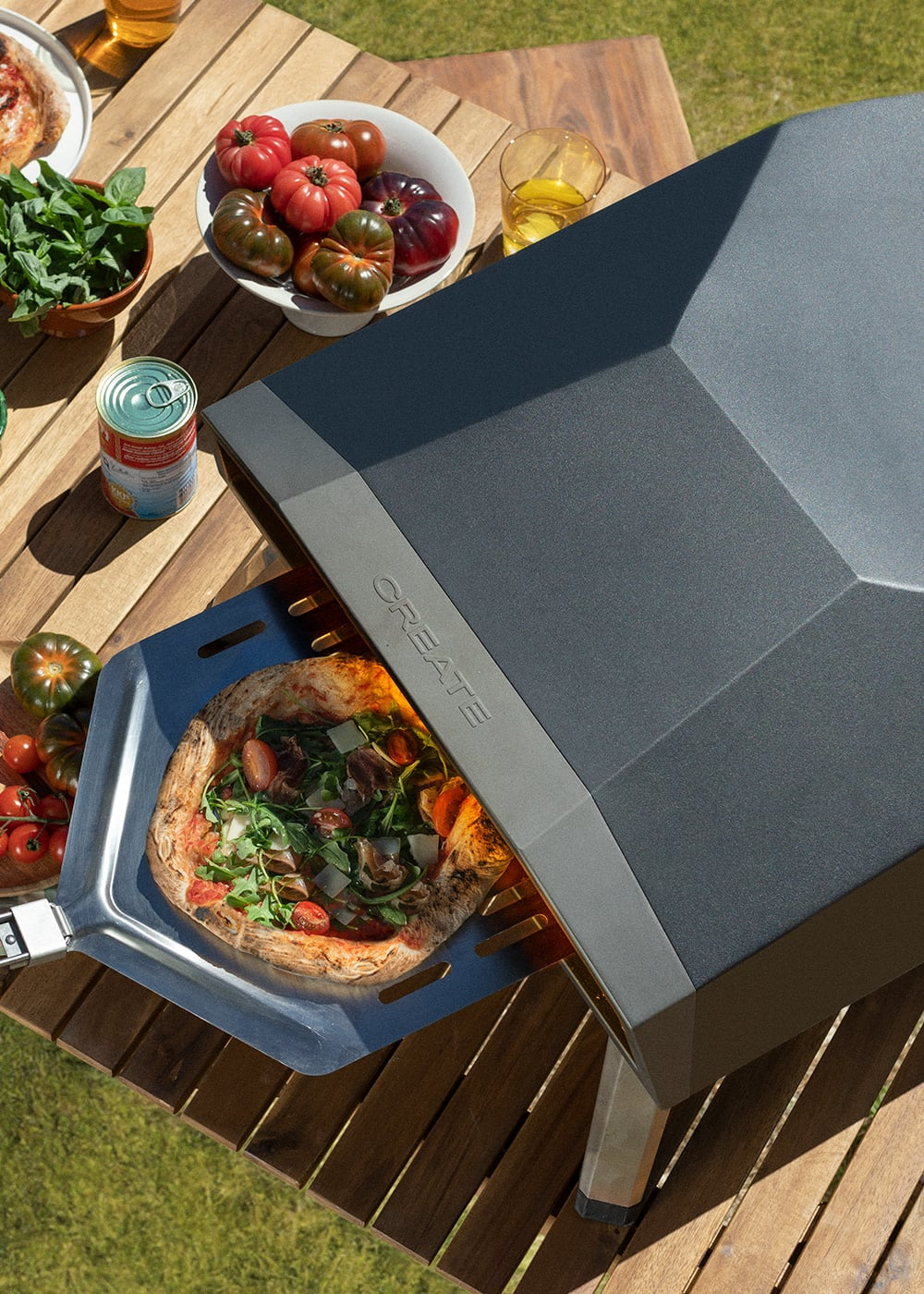 Una pizza se extrae de un horno de pizza gris oscuro a una pala metálica. Hay tomates y verduras cerca.