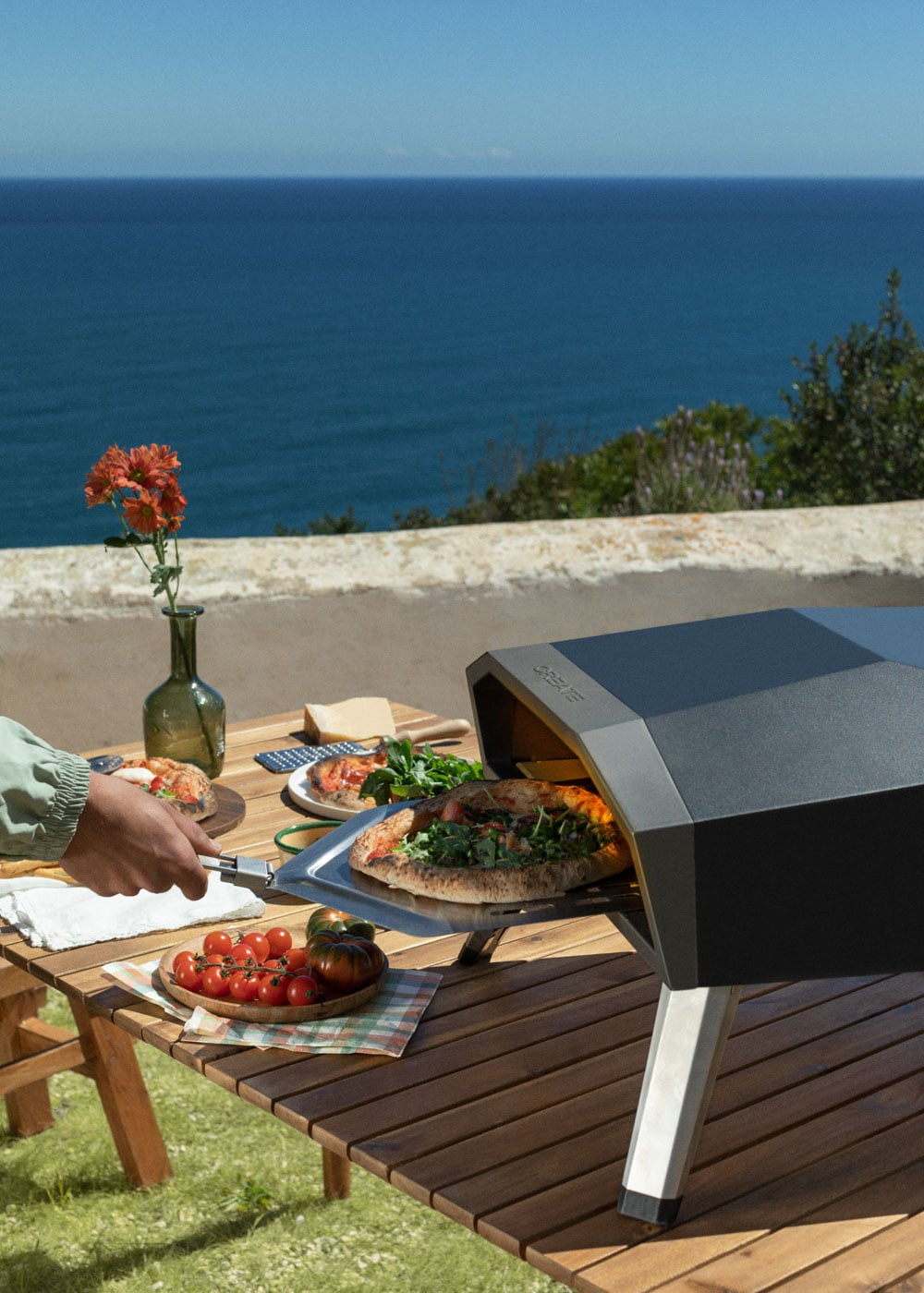 Preparación de pizza al aire libre con vista al mar. Mesa con pizzas, tomates y horno.