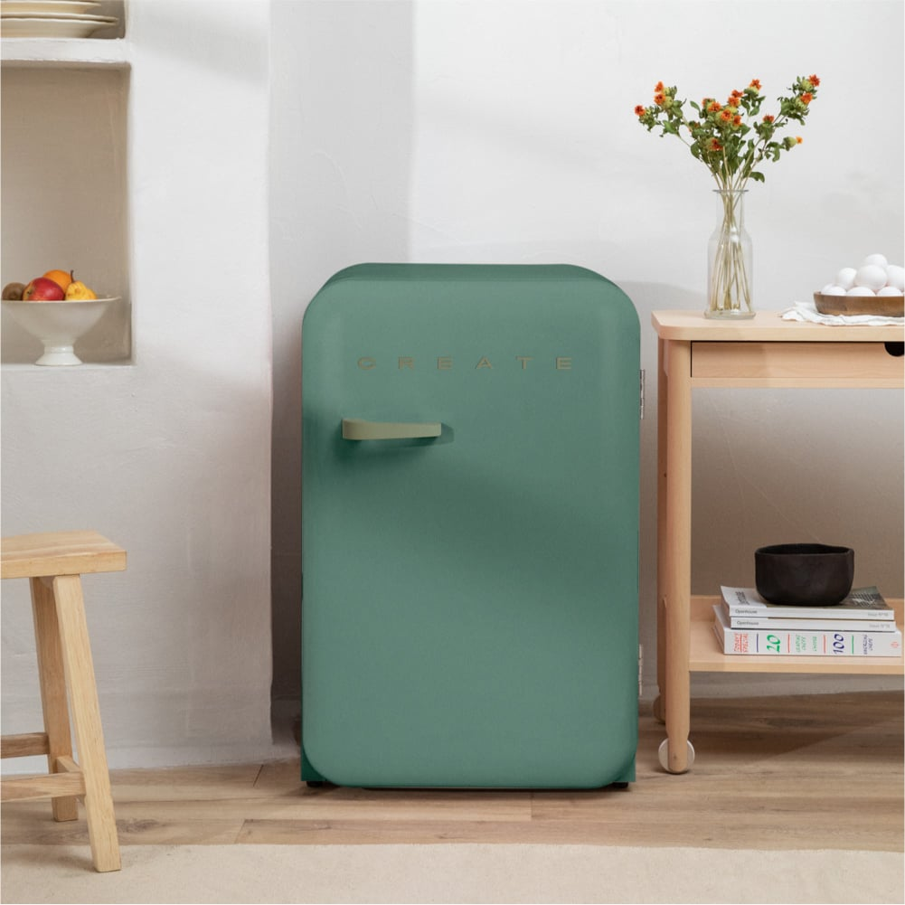Un refrigerador verde con la inscripción CREATE se encuentra junto a una mesa de madera con flores y huevos.