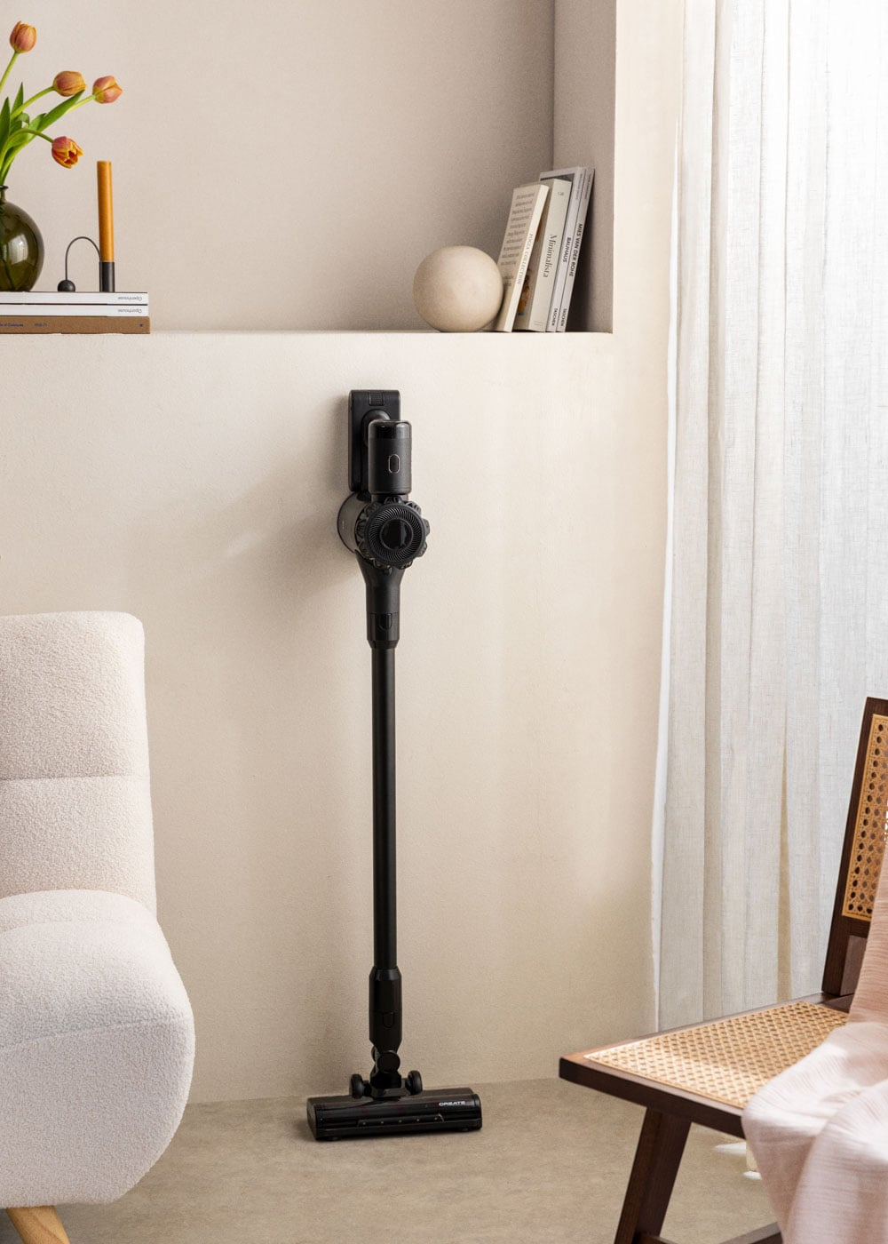 Aspiradora inalámbrica negra en soporte de pared, junto a un sillón blanco y silla de madera.
