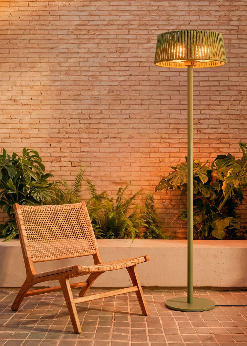 Una silla de madera y una lámpara de pie verde en un patio pavimentado contra una pared de ladrillos.
