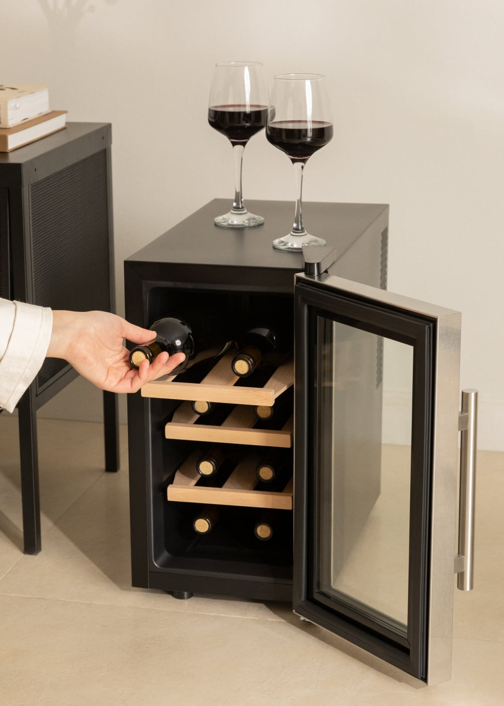Una mano coloca una botella de vino en una vinoteca abierta. Dos copas de vino tinto encima.