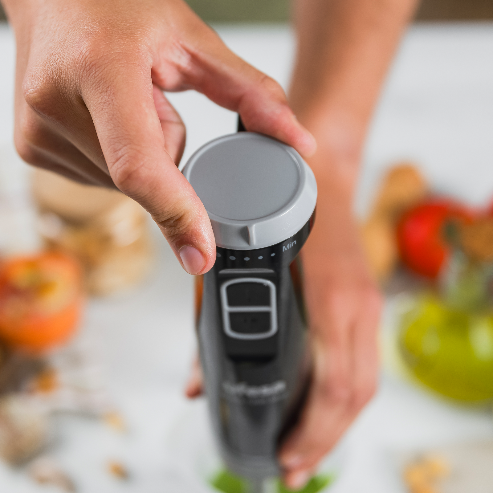 Mano che tiene frullatore a immersione nero con manopola e pulsanti. Ingredienti sullo sfondo.