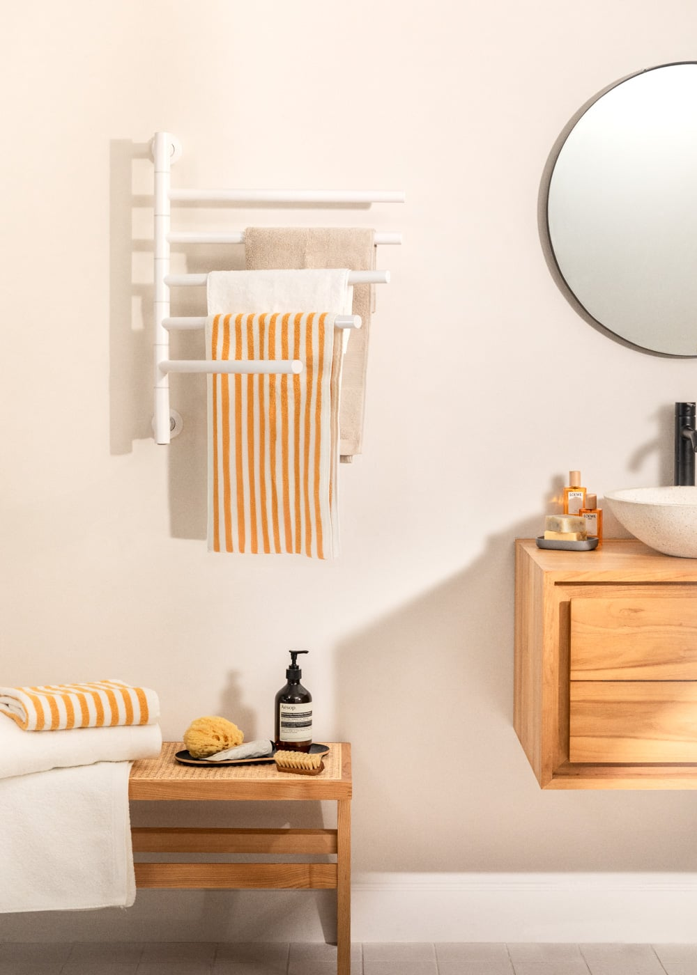 Una escena de baño con calentador de toallas, toallas, taburete de madera, jabón y lavabo.