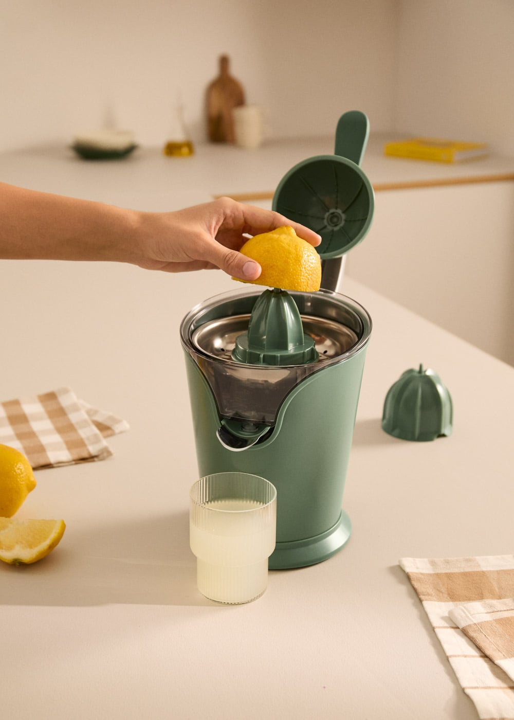 Exprimidor de cítricos verde con una mano sujetando medio limón. Un vaso de jugo al lado.