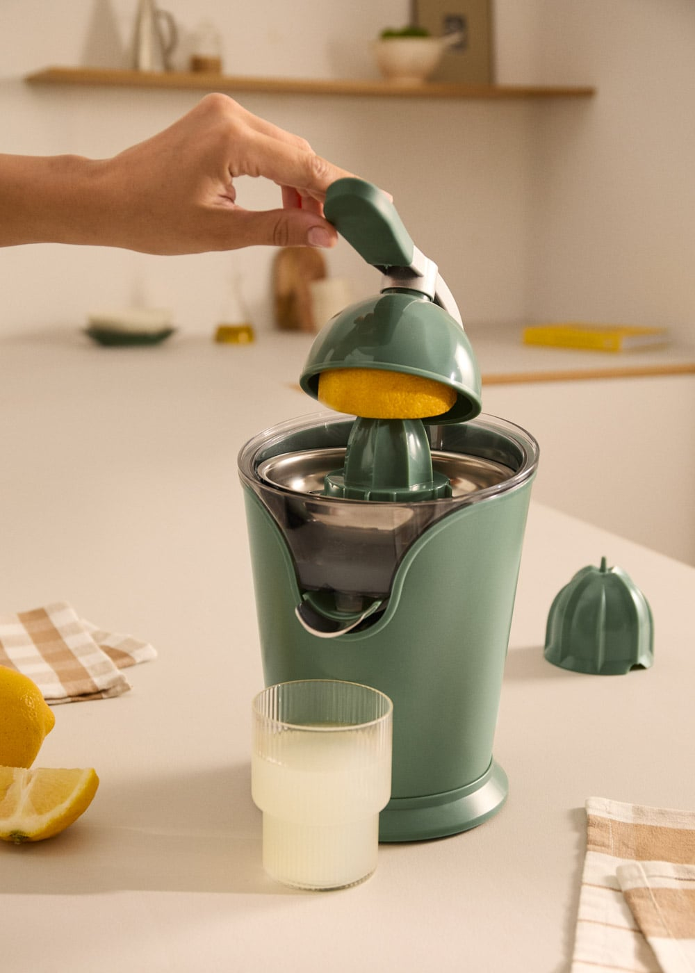 Una mano presiona media limón sobre un exprimidor verde. Al lado, un vaso de zumo.
