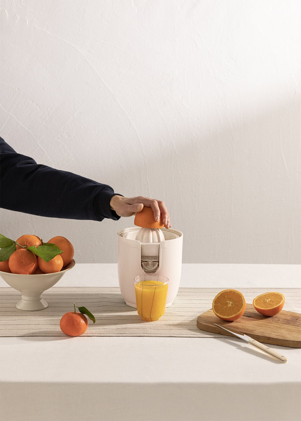 Una mano exprime una naranja en una licuadora. Al lado, un vaso de jugo y fruta.