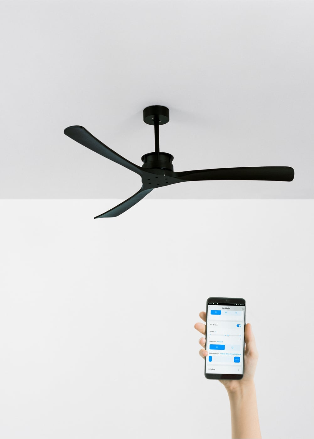 Un ventilador de techo negro con tres aspas está montado en un techo blanco. Una mano sostiene un teléfono inteligente con controles del ventilador.