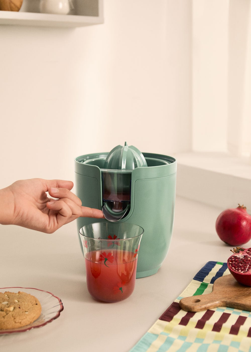 Una mano opera un exprimidor verde mientras el jugo fluye hacia un vaso. Hay granadas cerca.