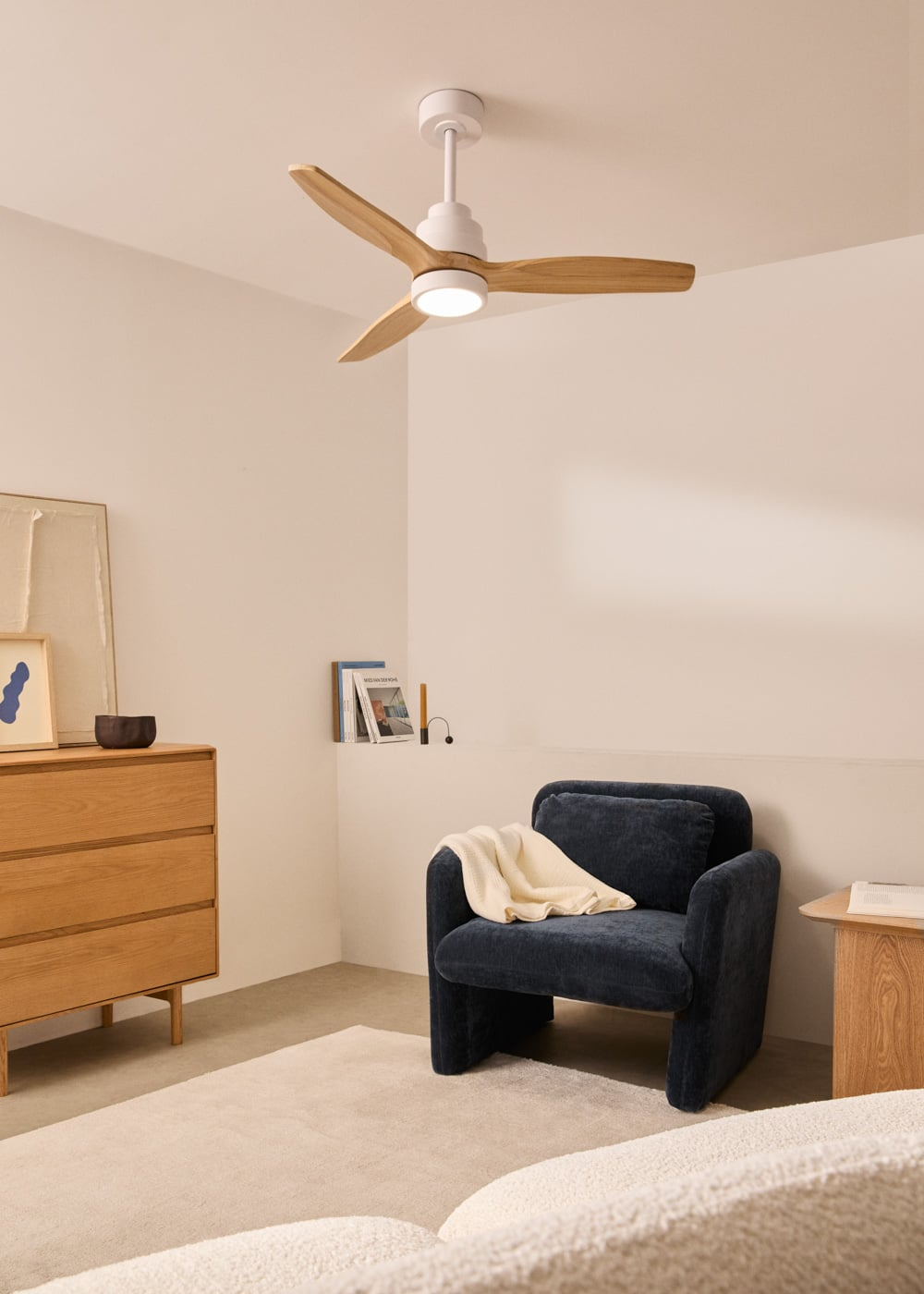 Un sillón oscuro con manta, cómoda de madera y ventilador de techo con luz.